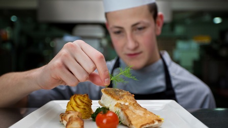 Frontale Aufnahme einer Person mit Schürze und Kochhaube, die auf einem Teller, auf dem ein Stück Fisch, Kartoffeln, Spinat und eine Tomate sind, ein Stück Dille legt