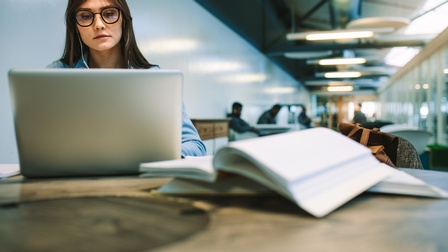 Person mit Brillen und Kopfhörern in den Ohren sitzt in Lesesaal und blickt auf aufgeklapptes Notebook vor ihr, am Tisch im Vordergrund verschwommen Bücher liegend