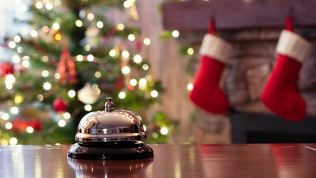 Eine Hotelklingel im Fokus, im Hintergrund verschwommen ein geschmückter Christbaum und Weihnachtsstrümpfe an einem Kamin hängend.