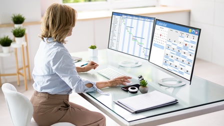 Person in Seitenansicht sitzt an Schreibtisch vor zwei Monitoren, auf denen Terminkalender zu sehen sind