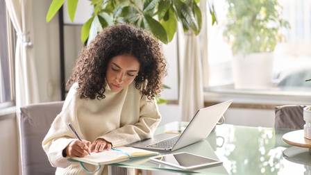 Eine Person sitzt seitlich an einem Glastisch. Vor der Person ist ein aufgeklappter Laptop. Rechts daneben ist ein aufgeklapptes Notizbuch, in das die Person etwas schreibt. Dahinter liegt ein Tablet. Im Hintergrund sind Topfpflanzen und Fenster