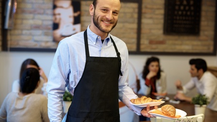 Lächelnde Person in schwarzer Schürze hält in einer Hand zwei Teller mit Croissants, im Hintergrund verschwommen weitere Personen an Tischen sitzend