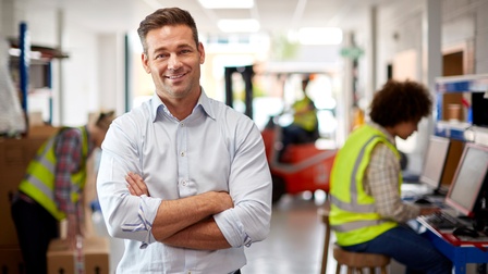 Halbporträt einer Person mit verschränkten Armen in der linken Bildhälfte. Im Hintergrund sind drei Personen, jeweils in Sicherheitswesten. Eine steht neben einem Karton. Eine sitzt hinter einem Bildschirm. Eine auf einem Gabelstapler