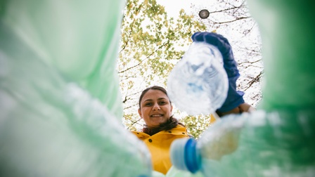 Person mit dunklen geschlossenen Haaren trägt Pet-Flaschen zusammen, Blick in die Kamera zwischen den Pet-Flaschen hindurch