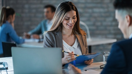Lächelnde Person blickt auf Clipboard mit Stift in der Hand, ringsum weitere Personen und aufgeklapptes Notebook