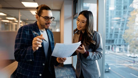 Zwei Personen stehen nebeneinander im Gang eines Büros. Die linke Person hält Zettel, auf die beide blicken