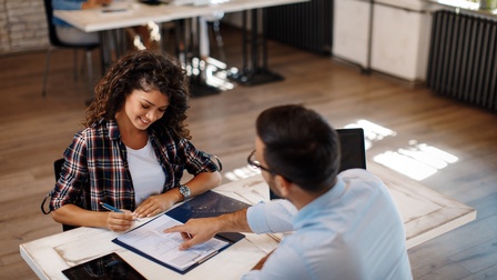Zwei Personen sitzen sich an Tisch in Büro gegenüber, eine Person hält Stift in Händen und blickt auf vor ihr liegendes Dokument, andere Person deutet mit Hand an Stelle in Dokument