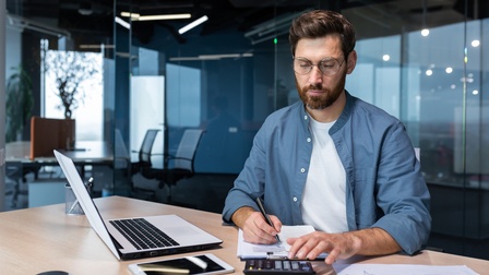 Eine Person tippt in einen Taschenrechner und macht sich Notizen. Sie sitzt auf einem Holztisch in einem Büro, neben ihr liegt ein Dokument mit einer Statistik, ein Tablet und ein Laptop. 