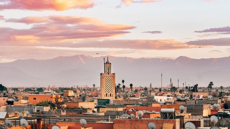 Panoramaaufnahme Marrakeschs mit den Dächern der Häuser sowie einem Minarett. Im Hintergrund ist die Gebirgsgruppe Atlas