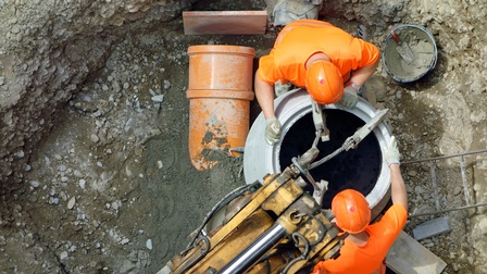Zwei Personen in oranger Schutz- und Arbeitsbekleidung mit orangem Schutzhelm führen Tiefbauarbeiten durch, Blick von oben