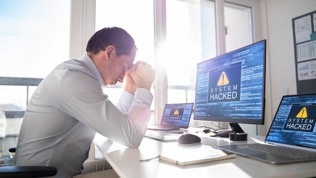 Eine Person sitzt am Arbeitstisch im Büro und hält die Arme auf dem Tisch gestützt vor die Stirn. Auf den Bildschirmen eines Computers und zwei Laptops erscheinen Cyber-Angriff-Symbole mit dem Schriftzug System hacked.