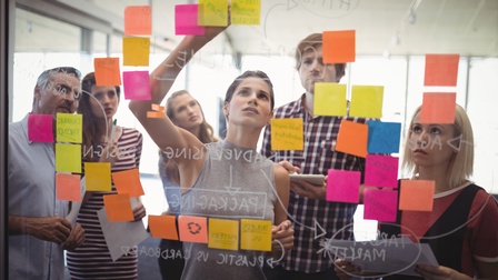 Mehrere Personen blicken auf transparentes Board auf dem bunte Post-its kleben, eine Person beschriftet Post-it