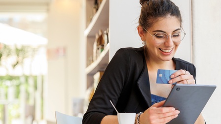 Lächelnde Person mit Brillen sitzt an Tisch, hält in einer Hand Bankomatkarte, in anderer Tablet, auf das sie blickt, vor ihr stehend weiße Tasse, im Hintergrund verschwommen Bücherregal