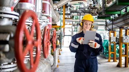 Eine Person mit gelbem Helm, Ohrenschützern, Arbeitskleidung, Handschuhen und Tablet, auf das sie blickt, steht in einem asphaltierten Gang in einem hohen Raum mit Rohren, Leitungen und Industrieanlagen. Direkt links neben ihr sind drei rote Radventile.