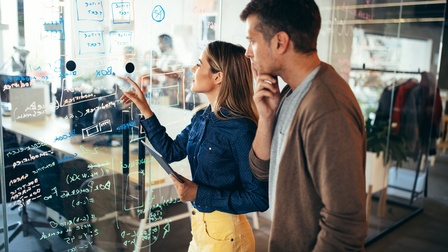 Zwei Personen stehen vor transparenter Wand, auf der Formeln und Schriften abgebildet sind, hinter der Wand verschwommen Großraumbüro