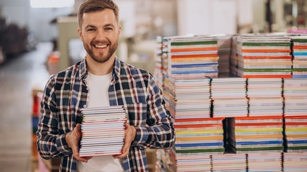 Porträt einer lachenden Person mit Bart und einem Stapel Büchern in beiden Händen. Hinter der Person ist ein hoher und breiter Stapel verschiedener, bunter Bücher