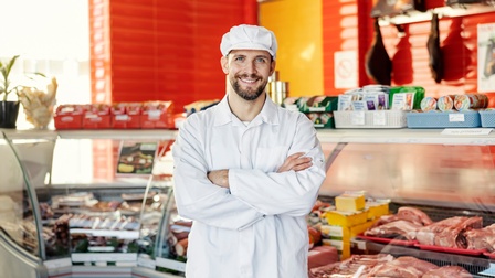 Porträt einer Person in weißer Arbeitskleidung mit verschränkten Händen vor einer Kühltheke mit verschiedenen Fleischstücken stehend. Auf der Theke liegen weitere unterschiedliche Produkte. An der Wand hinter der Theke hängen große Fleischstücke