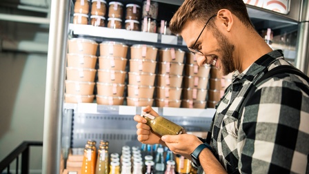 Eine Person mit Brille steht vor einem Kühlregal und hält ein kleines Fläschchen, das mit einer grünlichen Flüssigkeit befüllt ist. Im Kühlregal sind weitere Glasflaschen sowie rundliche Kartons mit Deckel