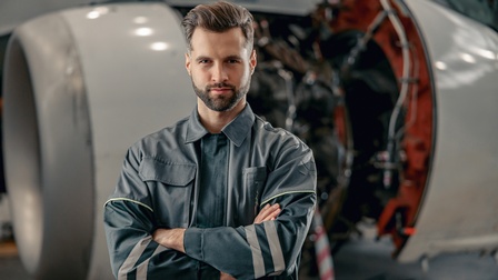 Porträt einer Person mit verschränkten Armen in Arbeitsoverall, im Hintergrund verschwommen Flugzeugturbine mit offen gelegter Technik
