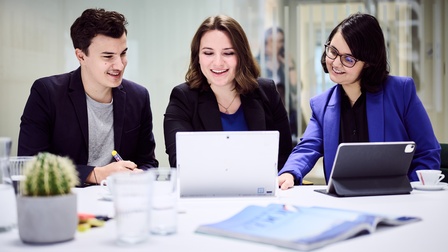 Personen sitzen an einem Tisch, vor ihnen sind Tablets aufgebaut, in eines schauen sie hinein. Auf dem Tisch sind Gläser, eine Karaffe, ein Magazin, Tassen und ein Kaktus platziert. Im Hintergrund ist eine Glaswand, in der eine Person gespiegelt ist. 
