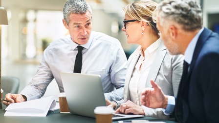 An einem Tisch sitzen drei Personen in Geschäftskleidung nebeneinander. Eine Person hat einen Laptop vor sich, eine ein Klemmbrett, die dritte ein Notizbuch. Vor jeder Person steht ein Kaffeebecher