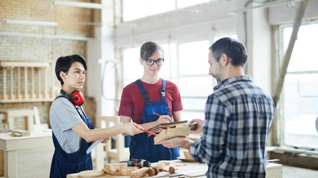 Drei Personen stehen um einen Werktisch in einer großen Werkstatt. Eine der Personen hält ein Holzstück und zeigt es den anderen beiden. Die anderen Personen halten jeweils einen Block und einen Stift