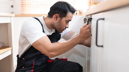 Seitliche Aufnahme einer Person mit Latzhose und kurzen, dunklen Haaren, die neben einem geöffneten Küchenunterschrank hockt und hinein blickt