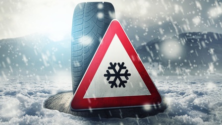 Ein Autoreifen ist  auf einer schneebedeckten Straße aufgestellt. Vor dem Reifen ist ein dreieckiges Schild, auf dem eine Schneeflocke ist. Es herrscht Schneeregen, im Hintergrund ist eine Berglandschaft zu erkennen. 