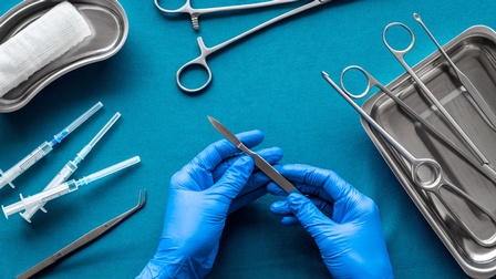 Auf einem türkischen Untergrund sind Werkzeuge für eine Operation platziert: ein Tuch, Spritzen, OP-Scheren, eine Pipette. Zwei Hände mit blauen Gummihandschuhen <br />
halten ein Skalpell. 
