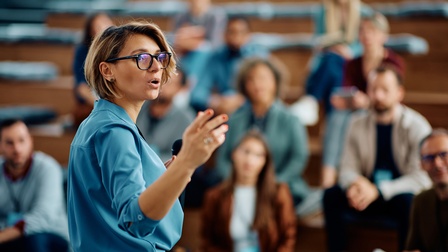 Leicht seitliche Aufnahme einer Person, die sich nach rechts aufdreht und spricht. In der linken Hand hält sie ein Mikrofon. Mit der rechten Hand deutet sie. Der Person gegenüber sitzen mehrere Personen in einem Hörsaal