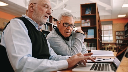 Zwei ältere Personen sitzen in einer Bibliothek am Tisch und blicken auf einen Laptopbildschirm, im Hintergrund stehen Bücher in Bücherregalen.