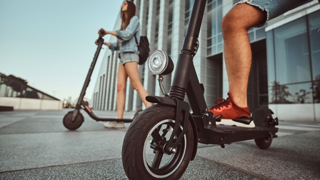 Detailaufnahme von einem Scooter, der von einer Person betrieben wird, im Hintergrund steht eine Person mit einem weiteren Scooter vor einem Gebäude auf einem Vorplatz