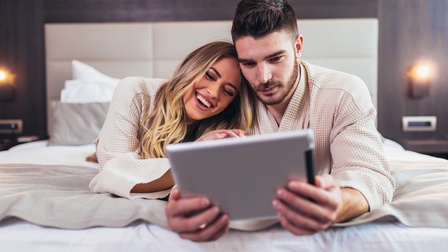Zwei Personen mit Bademantel liegen in einem Bett und blicken freudig auf ein Tablet