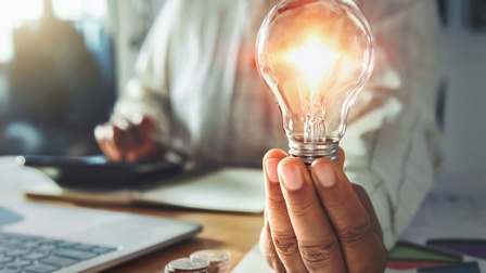 Die linke Hand einer Person hält mit den Fingern eine leuchtende Glühbirne. Die Hand ruht dabei mit dem Handrücken auf einem Tisch. Mit der rechten Hand tippt die Person in einen Taschenrechner. Vor ihr steht ein Laptop