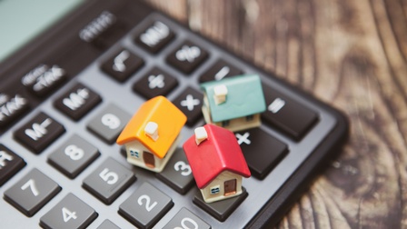 Nahaufnahme eines Taschenrechners, auf dem drei kleine Formen von Häusern stehen. Der Taschenrechner ist auf einem Holz-Untergrund platziert