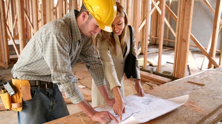 Zwei Personen stehen nebeneinander an einem Holzbrett auf einer Baustelle eines Gebäudes. Vor ihnen liegt ein Plan, auf den beide blicken. Die linke Person trägt einen Helm und einen Werkzeuggürtel