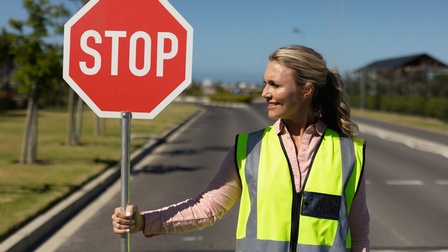Person mit langen blonden Haaren und gelber Warnweste steht auf einer Straße und hält ein Stoppschild in der Hand
