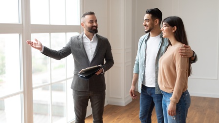 Drei Preisen stehen vor einer großen Fensterfront. Die Person links hält in der rechten Hand Unterlagen, blickt zu den beiden anderen und deutet aus dem Fenster. Die beiden anderen Personen stehen dicht nebeneinander und blicken aus dem Fenster