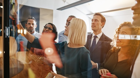 Seitliche Aufnahme von sieben Personen in Bürokleidung, die vor einer Glaswand stehen. Auf der Wand kleben viele verschiedene, bunte Post-its. Eine Person hat ihre Hand auf einem Post-it. Die anderen Personen blicken auf die Post-its