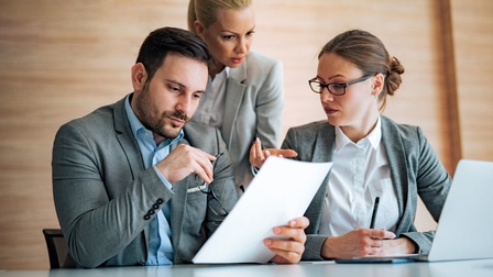 Zwei Personen in Bürokleidung sitzen nebeneinander an einem Tisch. Hinter ihnen steht eine weitere Person. Die links sitzende Person hält einen Zettel. Alle drei blicken auf den Zettel