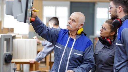 Drei Personen stehen seitlich neben einer hängenden Vorrichtung mit verschiedenen Knöpfen. Jede Person hat einen Gehörschutz um den Hals. Zwei tragen Schutzbrillen. Alle blicken auf die Gerätschaft