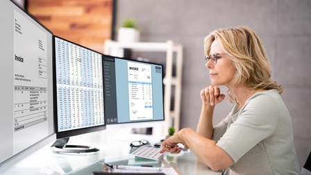 Person mit Brillen hält eine Hand an Kinn gestützt an Tisch sitzend, während sie auf Computermonitore mit Rechnungen und Zahlentabellen blickt