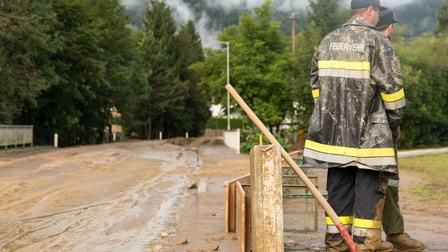 Zwei Personen in Rückenansicht in Schutzkleidung mit der Aufschrift Feuerwehr stehen neben wasserverschlammter Straße, die von Bäumen umsäumt ist, neben den Personen lehnt eine Schaufel an Holzbalken.