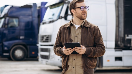 Frontale Aufnahme einer Person mit Brille, kurzen Haaren und Bart, die auf einem Parkplatz im Freien steht, ein Tablet hält und nach rechts blickt. Hinter der Person parken zwei Lastkraftwagen