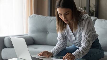 Person in gestreiftem Hemd arbeitet mit einem Laptop und rechnet mit einem Taschenrechner während sie auf einem Sofa sitzt