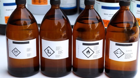 Braune Glasflaschen mit schwarzen Plastikverschlüssen nebeneinander gereiht, mit weißen quadratischen Etiketten und verschiedenen schwarzen Warnsymbolen: wie Totenkopf, toter Fisch und verdorrter Baum, Flamme und dergleichen