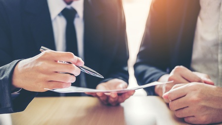 Frontale Nahaufnahme zweier an einem Tisch nebeneinander sitzender Personen in Anzügen. Die linke Person hält einen Zettel und einen Stift. Mit dem Stift deutet sie auf den Zettel