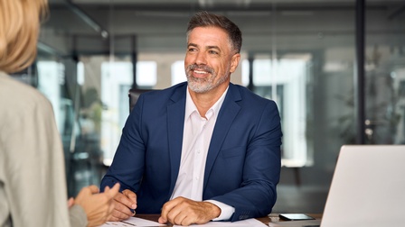 Frontale Aufnahme einer Person in einem Anzug, die an einem Tisch in einem Büro sitzt. Vor der Person liegen verschiedene Zettel. Ihr gegenüber sitzt eine weitere Person