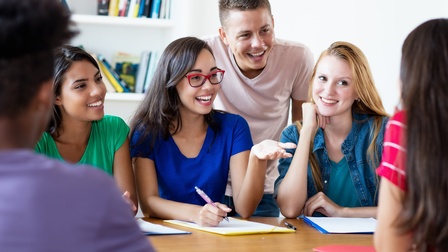 Eine Personengruppe sitzt gemeinsam an einem Tisch an der Uni und bereitet sich für Prüfungen vor.