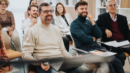 In mehreren Sesselreihen sitzen hintereinander mehrere Personen. Manche haben Zettel vor sich. Eine Person hat einen Laptop auf dem Schoß. Die meisten lächeln